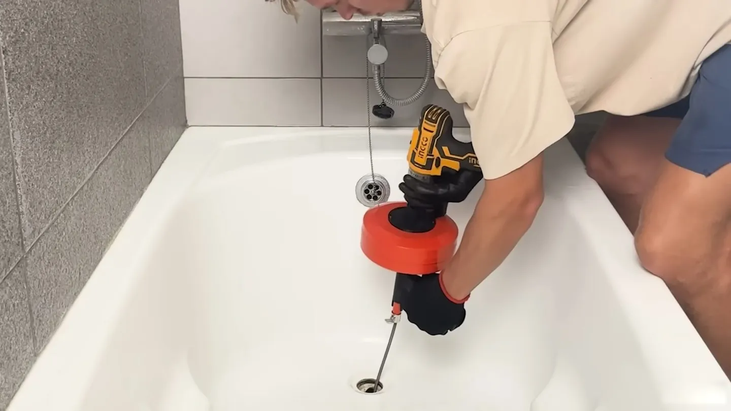 Person using a cordless drill with a red cable head at a bathtub drain
