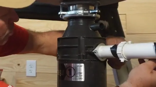 Black garbage disposal with a white discharge pipe attached by a clamp, a hand reaching in from the side