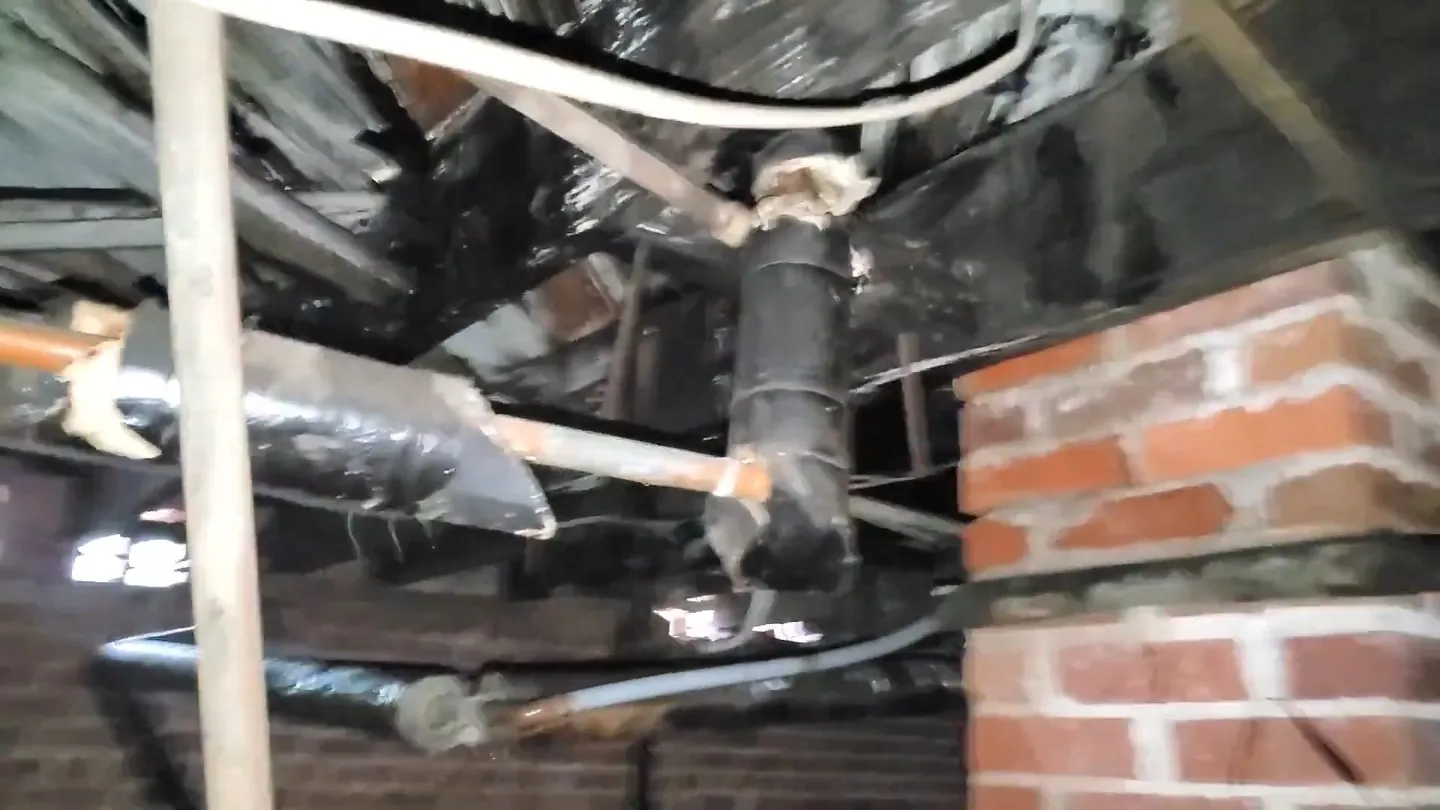Black insulated pipe and corroded fittings under a floor beside a brick wall