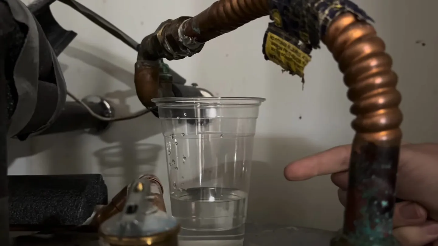 Clear plastic cup held under a copper water heater connection with dark insulation around the pipe