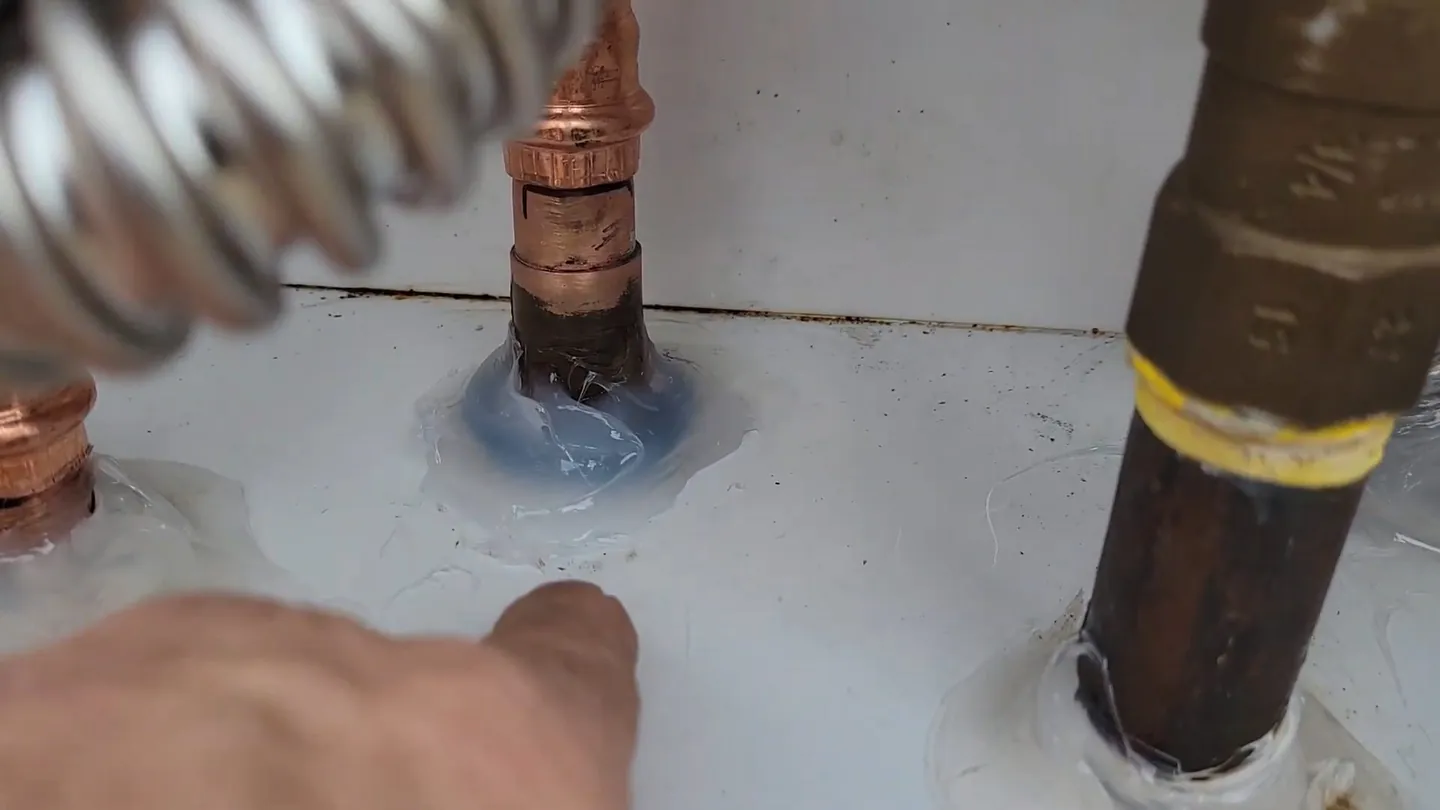 Hand pressing on a copper line beside a yellow-banded gas pipe at the top of a water heater