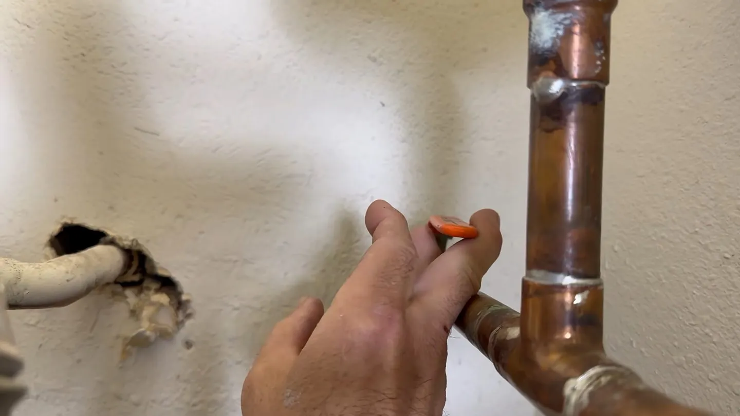 Hand holding a soldered copper pipe joint next to a rough hole in a wall where another pipe passes through