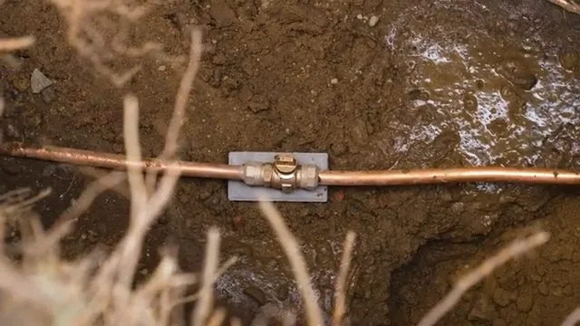 Copper water line running across an open dirt trench with a brass repair coupling in the middle and a flat metal plate beneath it