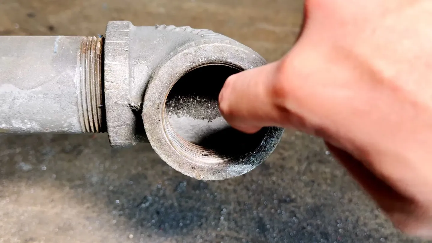 Fingertip at the open end of a galvanized pipe elbow where the cleanout cap has been removed