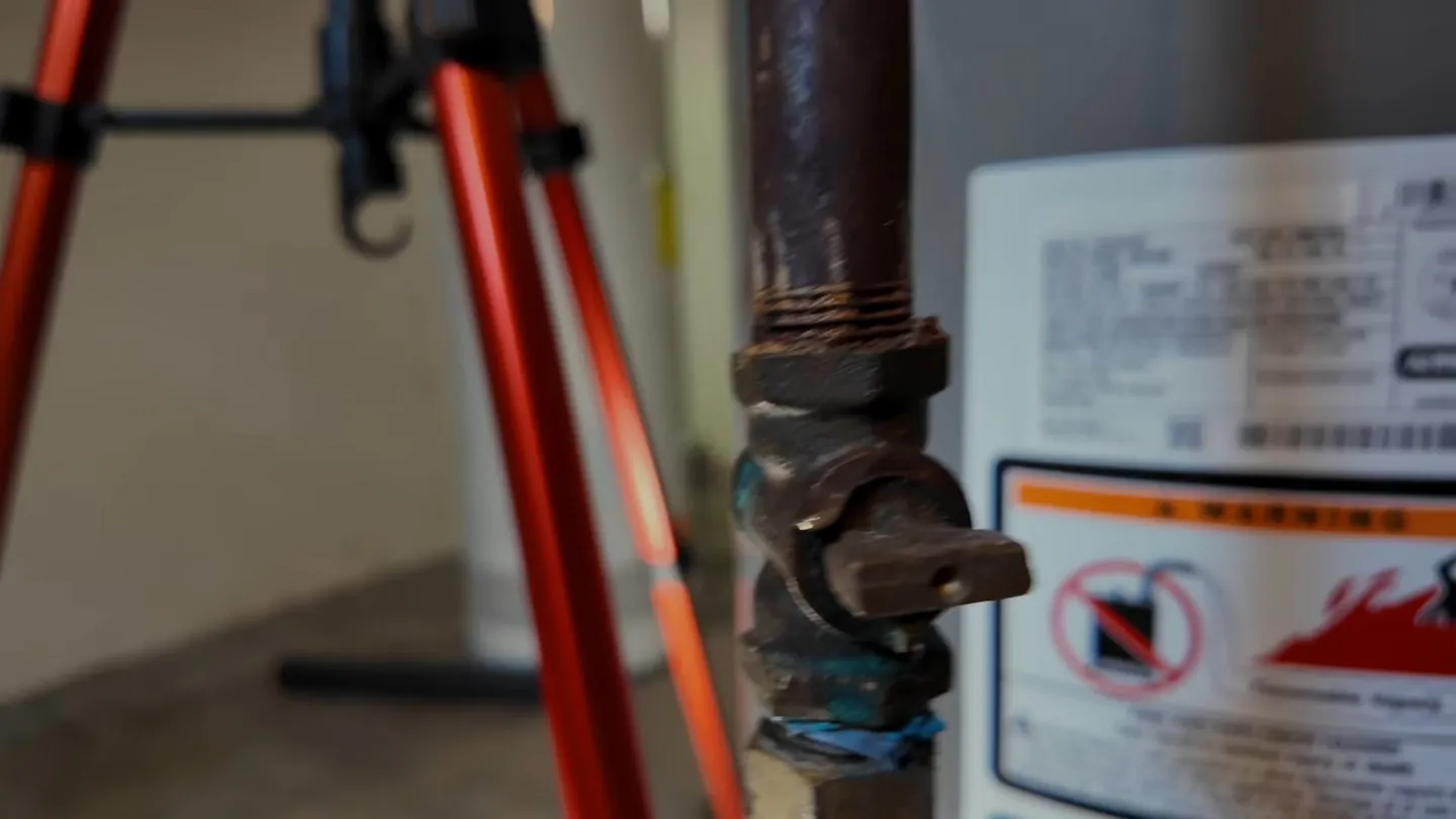 Black gas shutoff valve on an iron pipe beside a water heater, with an orange tripod behind it