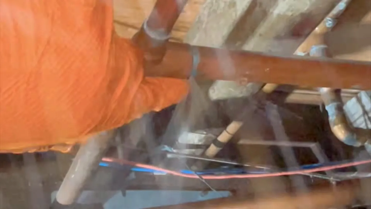 Orange-gloved hand gripping a copper pipe among floor joists with a fine spray of water across the frame