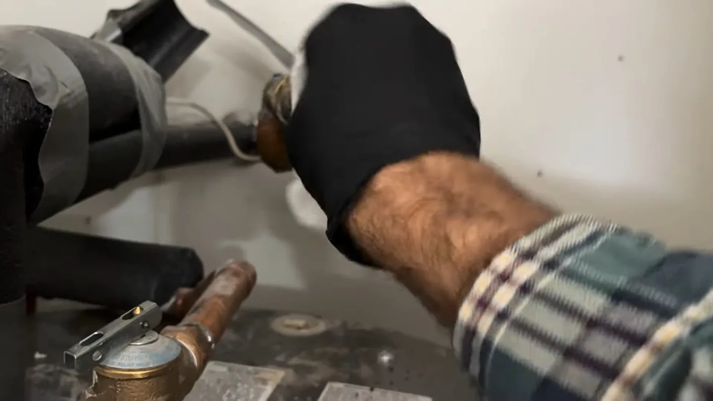 Gloved hand on a flexible hose connection above a water heater's copper valve, with a plaid sleeve in view