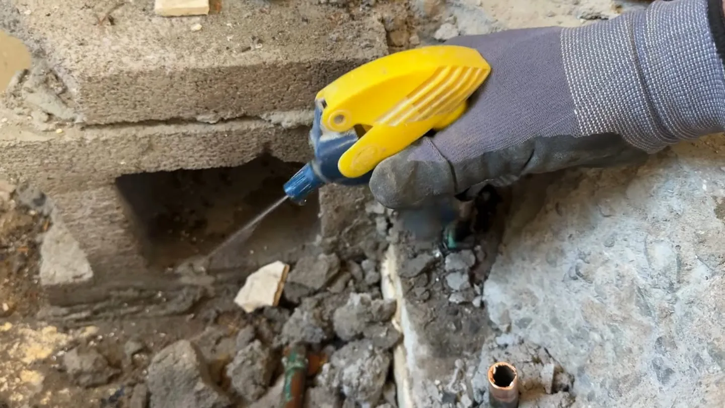 Gloved hand holding a yellow tool at a broken concrete slab opening beside a block wall with copper stubs