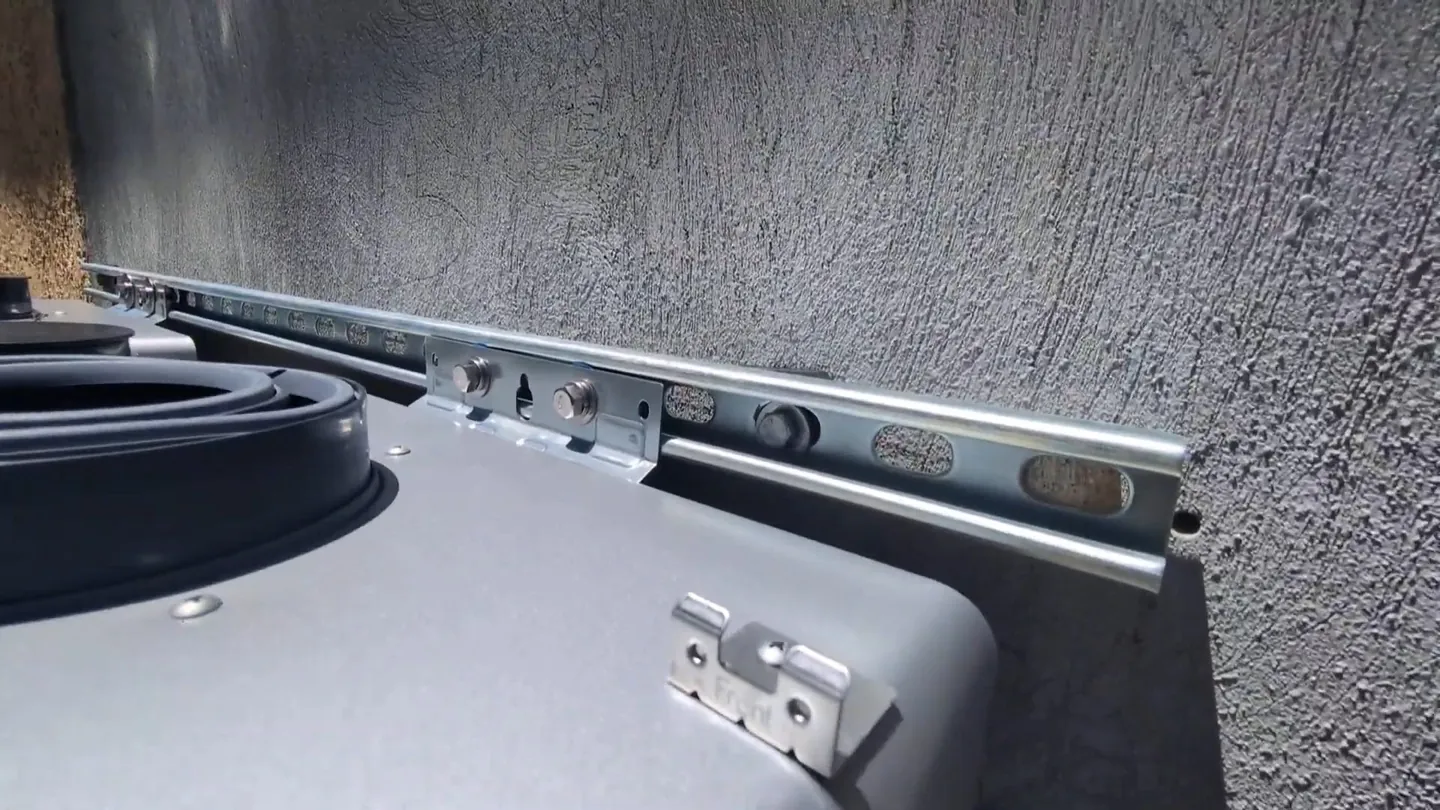 Gray appliance with a metal mounting rail and bracket against a textured stucco wall