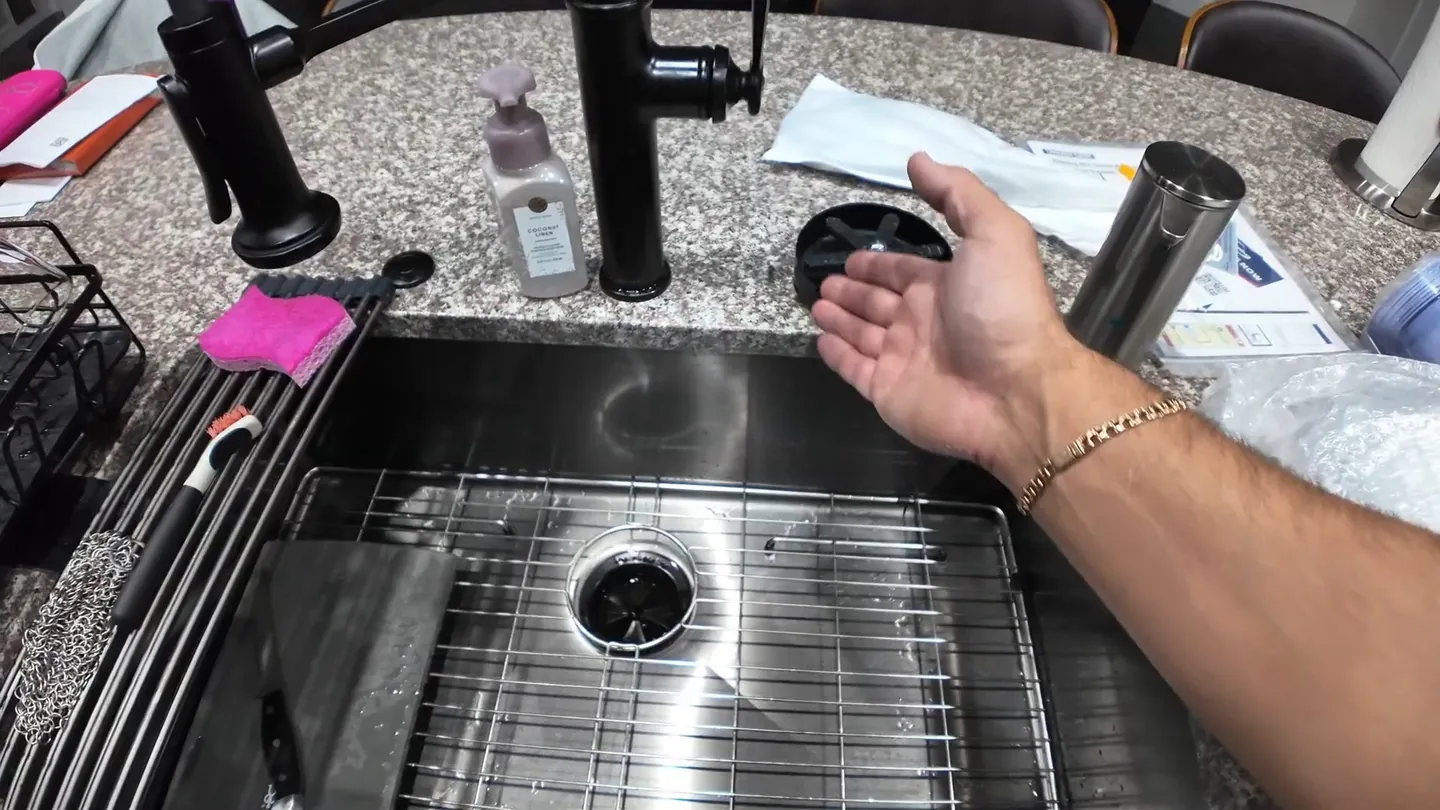 Hand reaching toward the drain of a stainless steel sink with a rack, a soap bottle and a black faucet