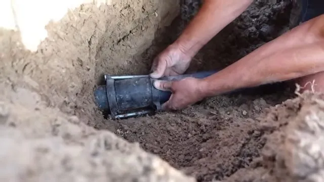 Two hands fitting a black coupling onto a pipe end at the bottom of a dirt trench