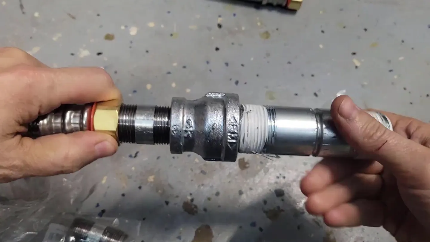Two hands joining a galvanized coupling between threaded pipe ends over a speckled floor
