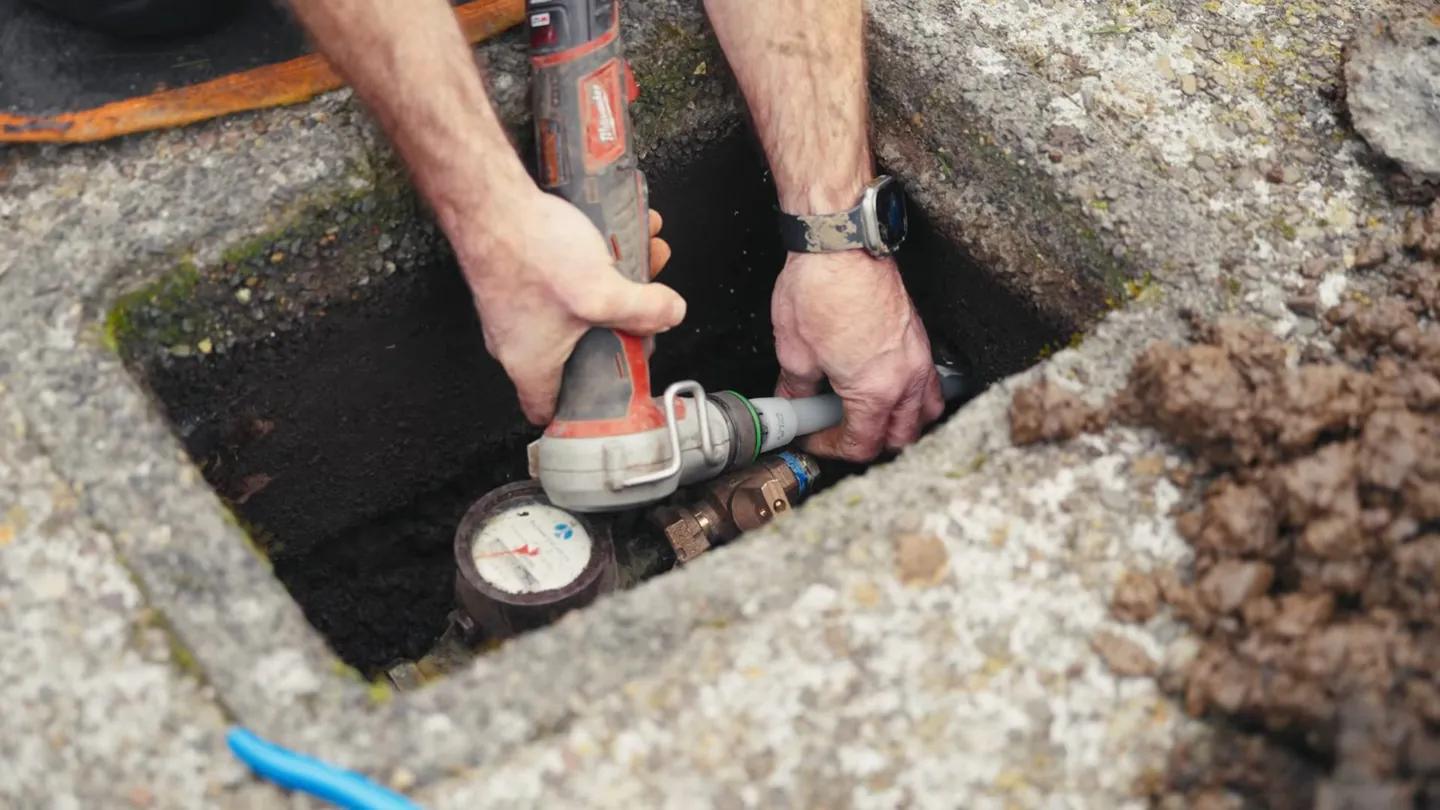 Hands holding a power tool against fittings inside a concrete meter box in the ground, with the meter at the bottom