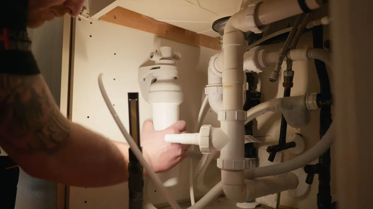Hands holding a filter housing in an under-sink cabinet crowded with white drain and supply pipes