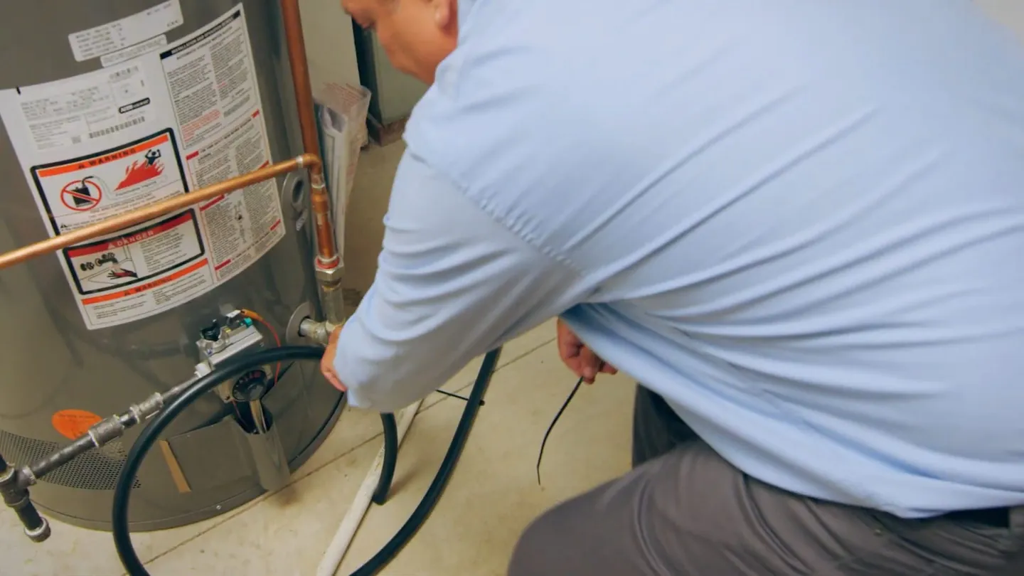 Man crouched beside a tank water heater with a green hose attached near its base