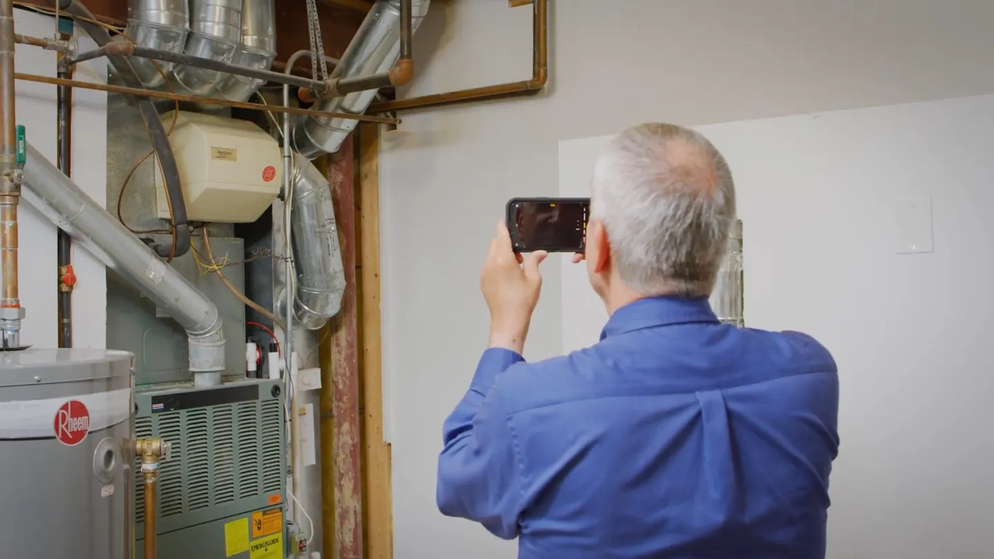 Man in a blue shirt photographing a wall in a utility room with a water heater and furnace beside it