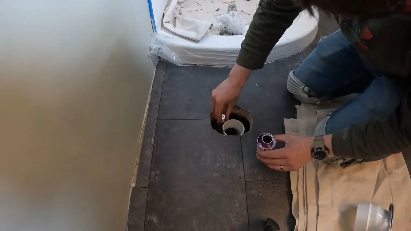 Person kneeling by an open floor drain on dark tile, holding a drain part and a small cup of purple liquid