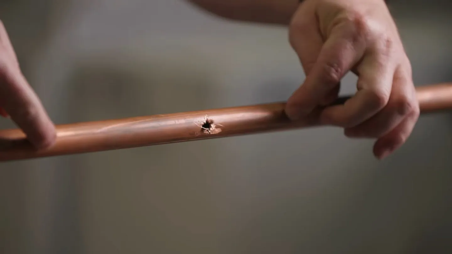Fingers holding a length of copper pipe with a small dark hole in its wall