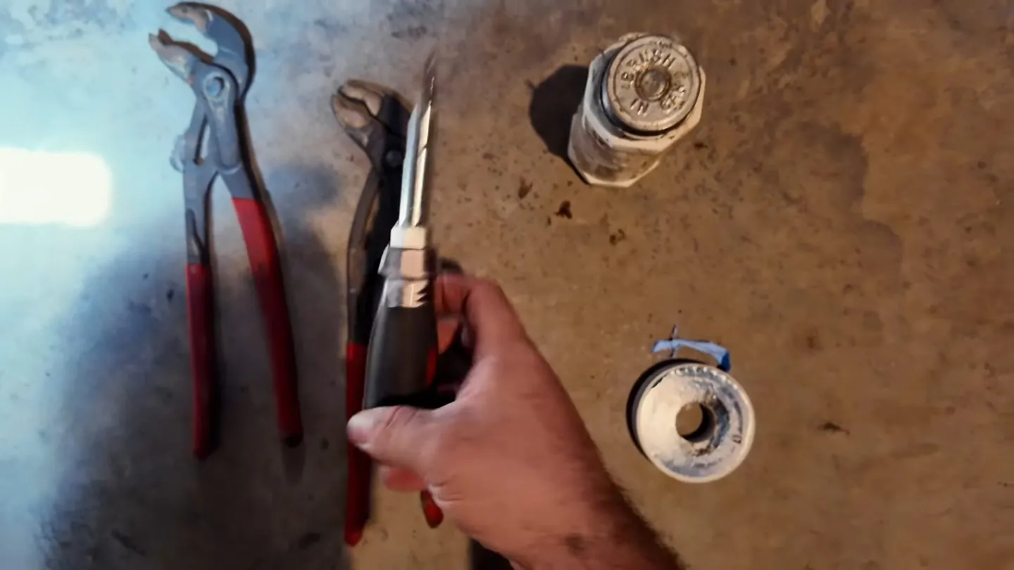 Hand holding pliers beside a pipe wrench, a threaded plug and a roll of thread tape on a worn surface
