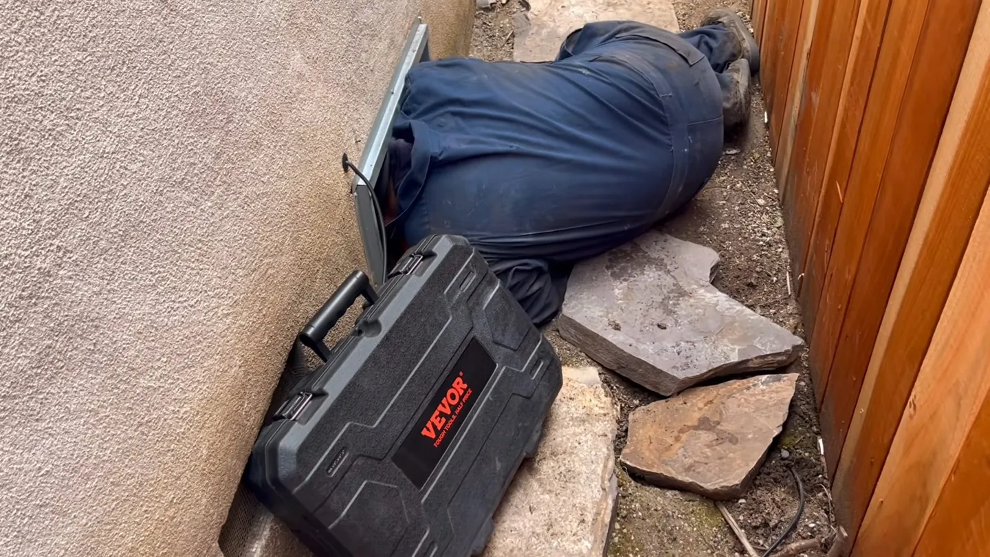 Plumber in blue coveralls lying on the ground beside a stucco wall, with a tool case next to him