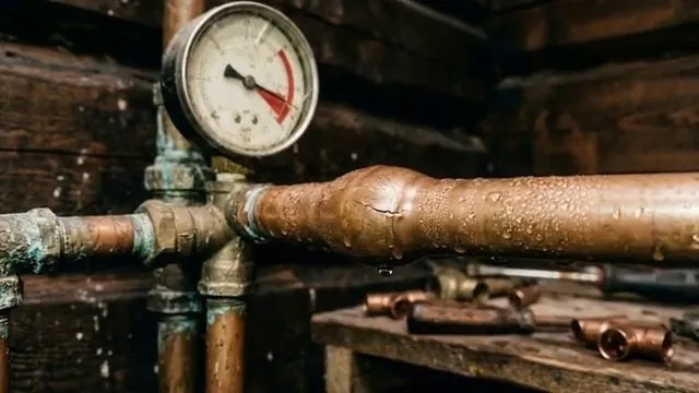 Pressure gauge fitted to copper supply pipes beside a large copper coupling, with drops of water on the pipe