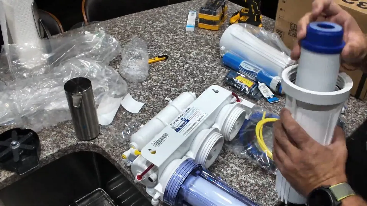 Reverse osmosis filter parts and packaging spread across a granite countertop while a hand lifts a filter housing