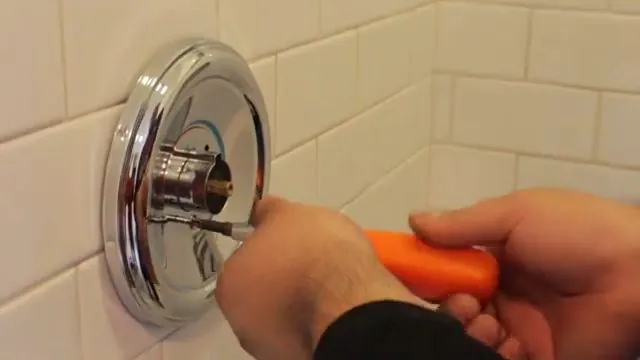 Orange screwdriver at the center of a chrome shower valve trim plate on a white tile wall