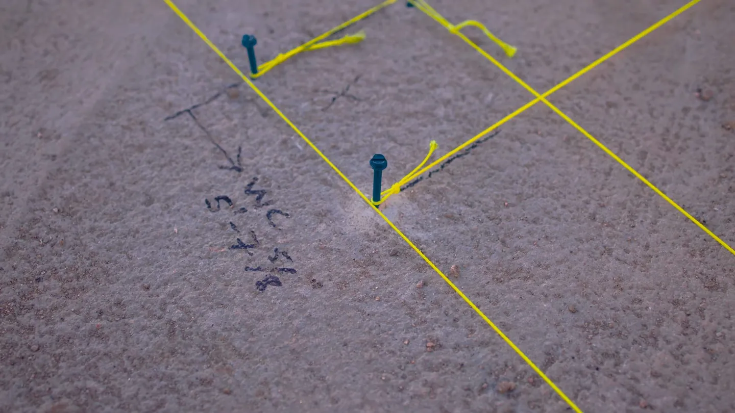 Yellow string lines and blue pins laid out on a concrete floor with a handwritten note beside one pin