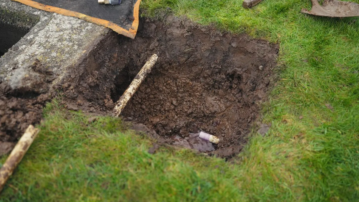 Small square hole dug in a lawn with soil piled beside it and a stick resting across one corner
