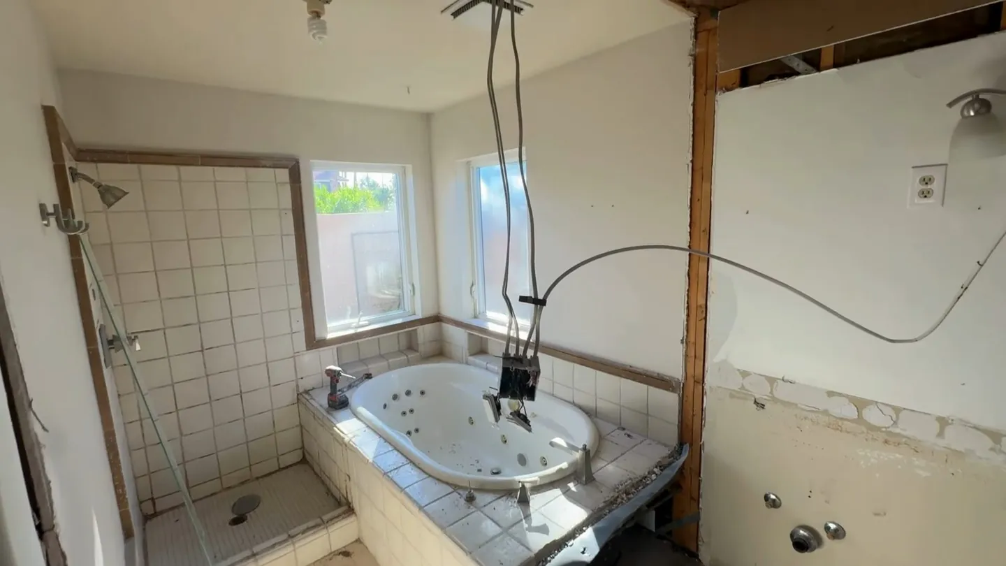 Bathroom stripped to studs with a tiled tub deck, a corner tub and wires hanging from the ceiling
