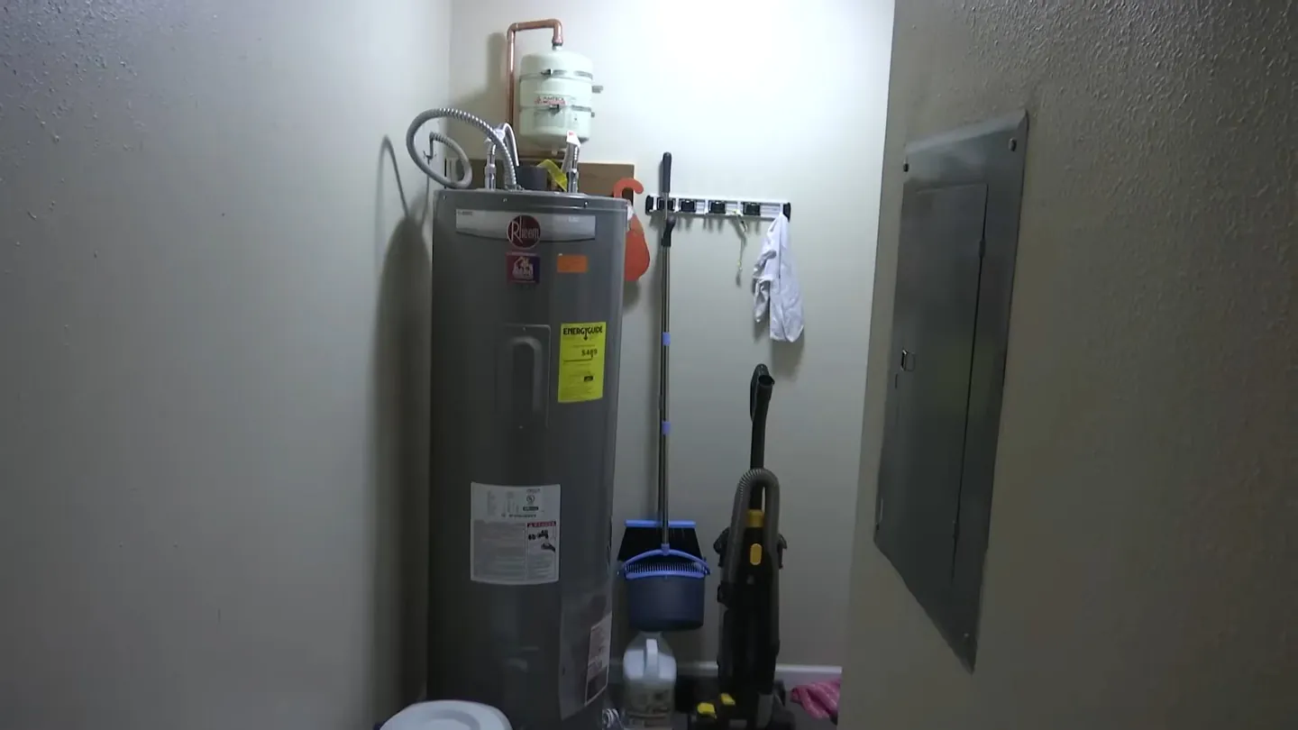 Tank water heater standing in a utility closet beside an electrical panel, with a mop and brush on the wall
