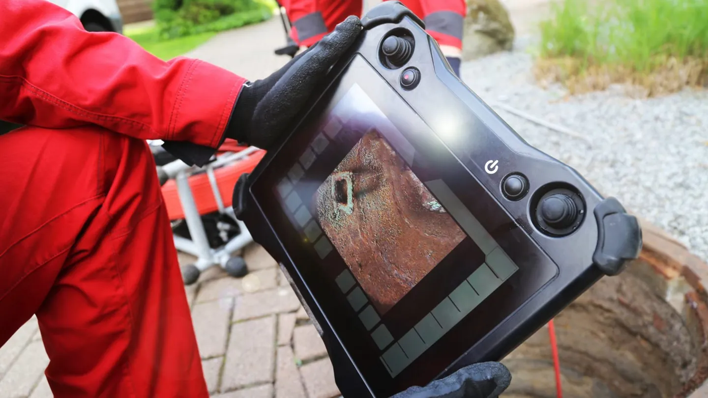 Technician in a red jacket holding a camera monitor with pipe footage on screen beside a paver walkway
