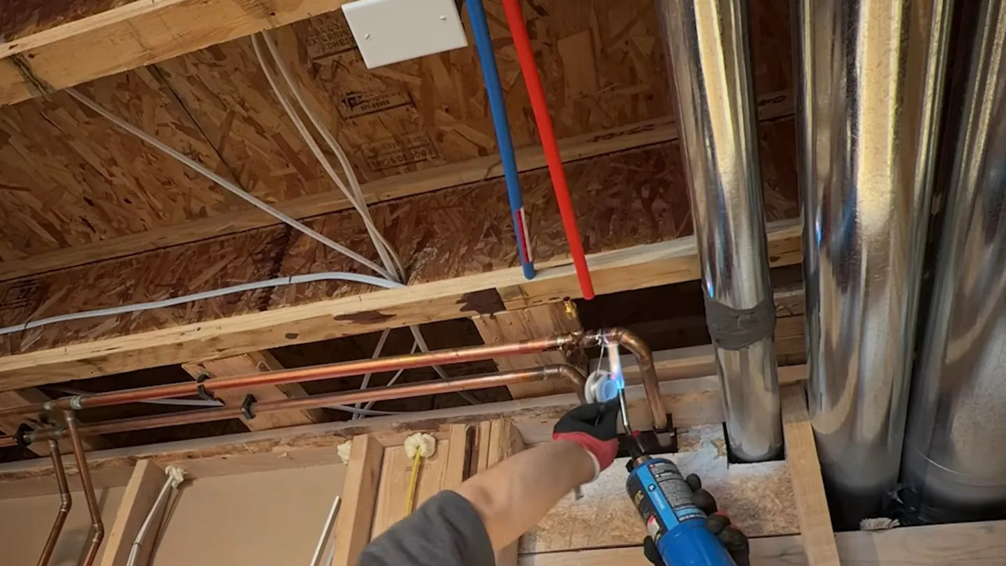 Technician with a handheld tool working in an open joist ceiling with red and blue supply lines and metal ducting
