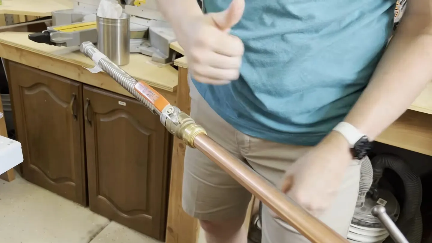Person in a teal shirt giving a thumbs up while holding a copper pipe fitted with a flexible connector and brass fitting