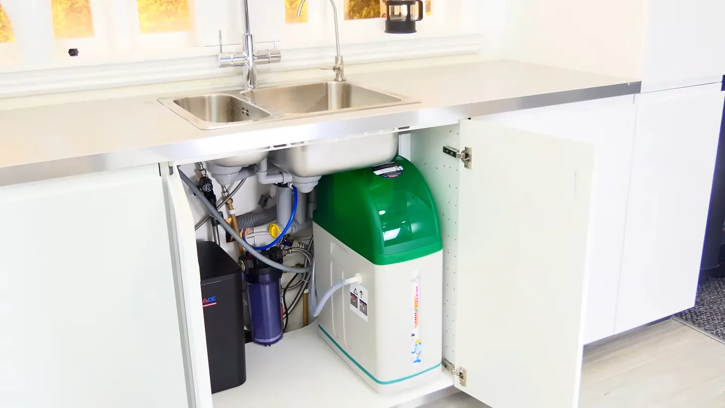 Open under-sink cabinet holding a green-topped softener and filter canisters beside the drain
