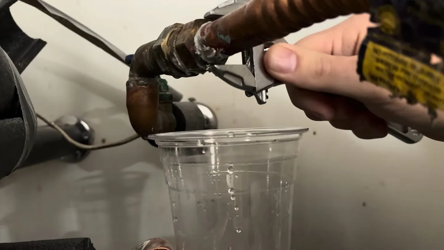 Hand at a water heater drain fitting with a clear plastic cup catching water below it
