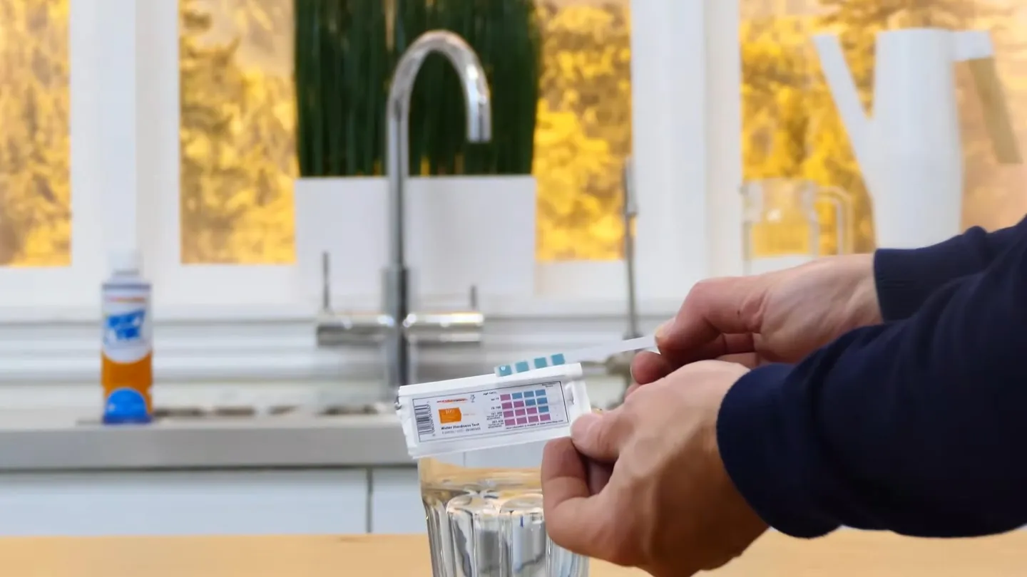 Hands holding a water test strip above a glass beside a kitchen faucet