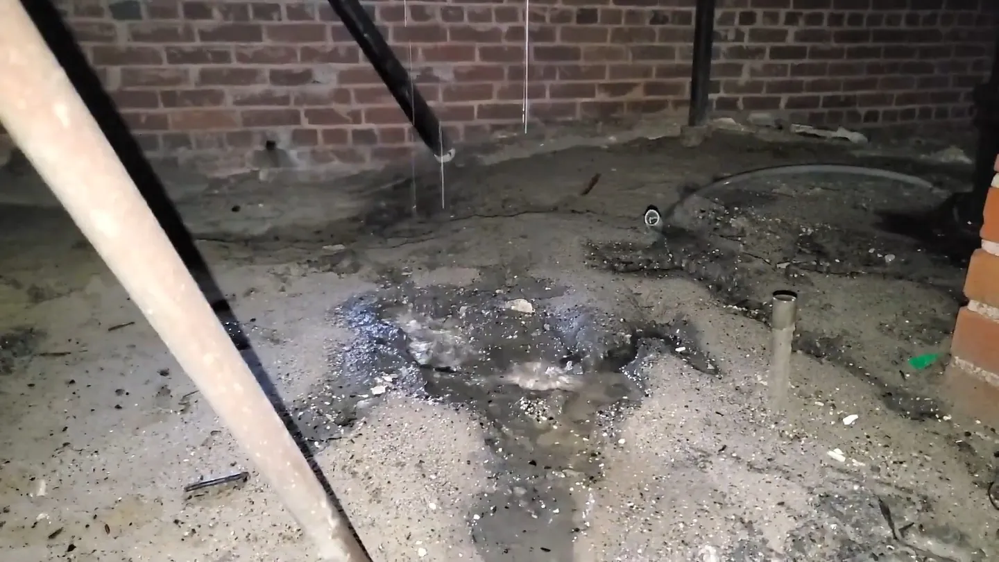 Wet dark ground with standing water beside a brick crawlspace wall and a pipe stub