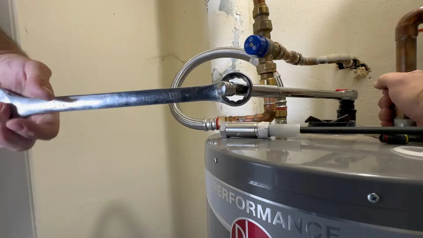 Hands holding a wrench on the anode rod at the top of a tank water heater, with a flexible line beside it